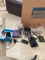Overview of the lot showing multiple electronic devices and accessories including Kindle, Nook, cameras, binoculars, calculator, cassette player, cables, and projection clock on floor.