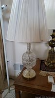 Full view of crystal lamp on wooden table showing off-white pleated fabric shade, crystal patterned lamp body, and gold-toned base.