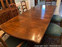Dining table with three removable leaves placed in the middle, photographed with six matching wooden ladder-back chairs including two with arms and four armless chairs, all with green cushioned seats. Tabletop protector present.