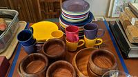 Overall view of stacked colorful dinner and salad plates with 7 mugs and wooden bowls including large and smaller salad bowls.