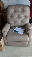 Front view of beige electric lift chair showing padded armrests, tufted backrest with buttons, wired remote, and instruction manual on seat.