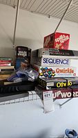 Photo showing stacked game boxes including Spill and Spell, Saitek Pro Bridge 310, Sequence, Qwirkle, Othello, Up Words, and Jenga.