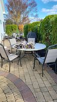 Full view of round patio table with transparent glass top, with four beige mesh chairs around it on a stone patio. Umbrellas and table cover placed on table.