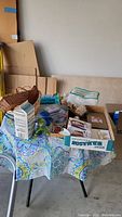 Overview of lot including assorted boxed and loose items on a table with tablecloth in garage setting.