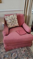 Front view of the red upholstered armchair with embroidered throw pillow and fabric arm covers, placed on a patterned rug near a window.