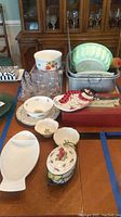 Photo displaying various ceramic serving bowls, casserole dishes, glass mugs, and metal cookware items arranged on a table.