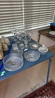 Blue table displaying several Lenox Windswept pattern wine glasses, plain wine glasses, Waterford bud vase, decorative glass cube, Mikasa bowl, candy dishes and covered sugar bowl.