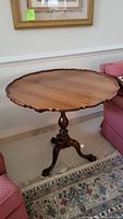 Full view of the round wooden tilt top table with scalloped pie crust edged top and carved pedestal base ending in claw feet.