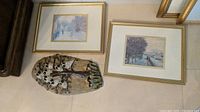 Two framed prints placed side by side with the pottery owl wall hanging in front, all on the floor with a beige background.