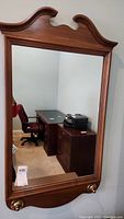 Full view of the wood-framed rectangular wall mirror with decorative curved top and brass-tone accents at bottom corners.