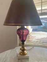 Full view of the lamp showing amethyst thumbprint glass body, brown lampshade with gold trim, brass fittings, and white marble base on a tabletop.