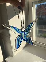 Photo showing hand holding the butterfly glass suncatcher by the hanging metal ball chain with a small key attached, sunlight shining through window