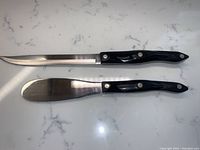 Two Cutco utensils shown side by side: a serrated knife and a spatula spreader, both with black handles.