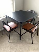 Photo of vinyl covered square folding table with three brown vinyl padded folding chairs around it, showing overall lot contents and condition.