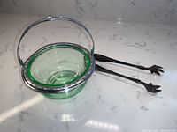 Green depression glass basket with detailed chrome metal frame and handle placed on white marble surface with silver plate claw foot ice tongs next to it.