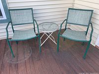 Two green metal frame patio chairs with woven mesh seats and backs next to a small round folding table with a glass top, all placed against the exterior siding of a house on a wooden deck.