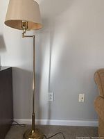 Full view of the brass swing arm floor lamp next to wall showing beige shade and straight pole.