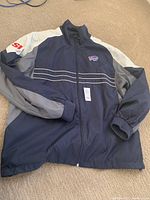 Full view of navy, gray, and white windbreaker jacket with Buffalo Bills logo and SI logo on sleeve laid flat on carpet.