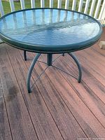 Round outdoor table with textured glass top and metal frame shown from an angle on a wooden porch floor.