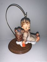 Side view of signed Hummel figurine sitting on a wooden base stand. Angelic child on a cloud holding an orange item with a basket on back.