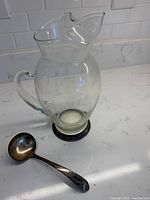Clear hand blown glass pitcher with sterling silver rim base next to small sterling silver ladle on white surface.
