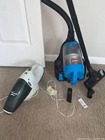Photo showing the Bissell Zing canister vacuum and the Black and Decker handheld vacuum with charging cable and accessories on carpeted floor.