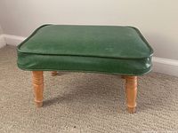 Footstool showing green vinyl padded seat top with minor surface wear and turned wooden legs on carpeted floor.