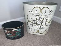 Two metal waste bins side by side on carpet. Left smaller black bin with horse painting, right larger white bin with black and gold scroll design.