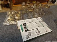 Photo of eleven clear glass jars arranged on carpet and multiple gold-colored metal rings and lids in plastic bag and box.