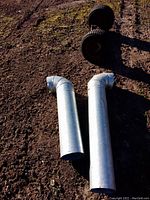 Two galvanized metal heat pipes with elbow joints lying on soil, one longer than the other.