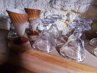 Two cone-shaped glass holders designed to resemble waffle ice cream cones with brown bases and white interiors, displayed upright on a wooden surface next to glass ice cream cups.