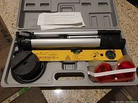 Complete laser level kit shown inside open gray plastic carry case. Kit includes adjustable aluminum tripod, yellow laser level tool, two red circular timer lights with translucent covers connected by white strap, and instruction sheet.