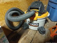 Ridgid shop vac shows the body with attached black flexible hose, placed on wooden floor board surface.
