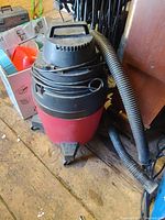 Red and black Shop Vac wet/dry vacuum cleaner sitting on a wooden floor with an attached gray hose coiled around the unit. Shows signs of use.