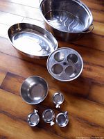 Overview of all items in the lot: large pan, tray, egg poacher, bowl, and clip-on cups on wooden floor.