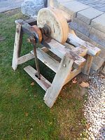 Side angle view of vintage foot operated grinding stone wheel mounted on an aged wooden frame, showing pedal and wheel condition.