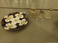 Full view of the decorative porcelain plate with floral design alongside three vintage glass shakers of different shapes and sizes.