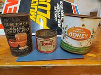 Overview of three vintage tin cans stacked on a table including Heinz Tomato Juice, Toddy, and Pure Canadian Honey cans with visible wear and rust
