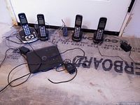 Photo showing four wireless cordless home phone handsets arranged in a row with their charging bases, connected wiring, and one main base station unit with a power adapter on a floor surface.