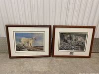 Two framed wildlife art pieces side by side, one of a polar bear and the other a black bear with cubs near a forest lake, both signed and matted.