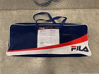 Blue, white, and red Fila carrying bag for badminton set, with handles, placed on floor.