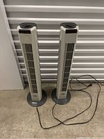 Two Hampton Bay tower fans standing side by side on a concrete floor, showing full height and form.