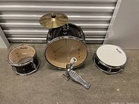 Photo showing bass drum with cymbal, pedal, and two snare drums on floor against a wall.