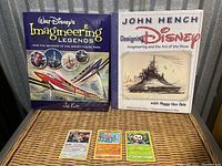 Two Disney Imagineering books and three sealed Pokémon cards displayed on wicker surface.