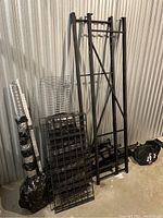 Disassembled black metal shelving parts leaning against a corrugated metal wall including black vertical supports, wire mesh shelves, and horizontal beams.