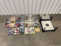 Photo showing an assortment of 14 Xbox 360 video game cases, a red WWE DVD disc, and a small WWE wrestling ring toy on a metal surface.