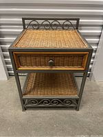 Front view of the bedside table showing woven wicker drawer, metal frame with decorative scrollwork on base and top back panel.