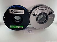 Two spools of 3D printer filament on a white background, one black with a blue PLA label, the other gray with Duramic 3D PETG label.