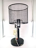 Front view of the lamp showing black mesh shade and round black base with lamp cord coiled in front.