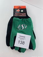 Pair of green and black CFL sport utility gloves with embroidered Roughriders 'S' logo on back, attached to black and orange branded packaging.